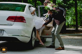 At a church in Budapest, Hungary, the groom gently assists the bride as she steps out of the car, bouquet in hand, her foot just about to touch the ground, marking the beginning of their special day together.